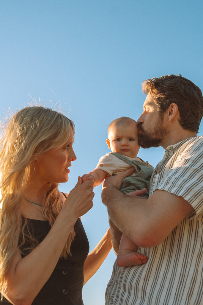Costa Mesa family photographer captures parents with baby at golden hour, dad kissing baby's head