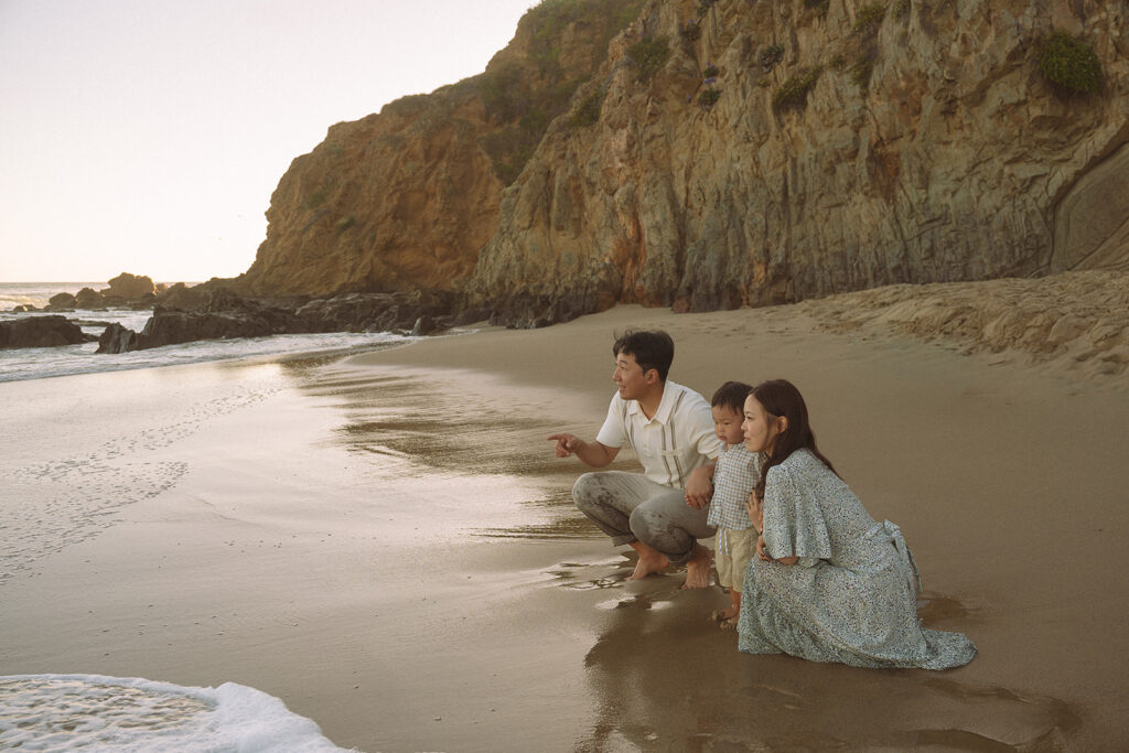 Family of three crouched at the shoreline looking out at the ocean during a Crescent Bay family session in Laguna Beach