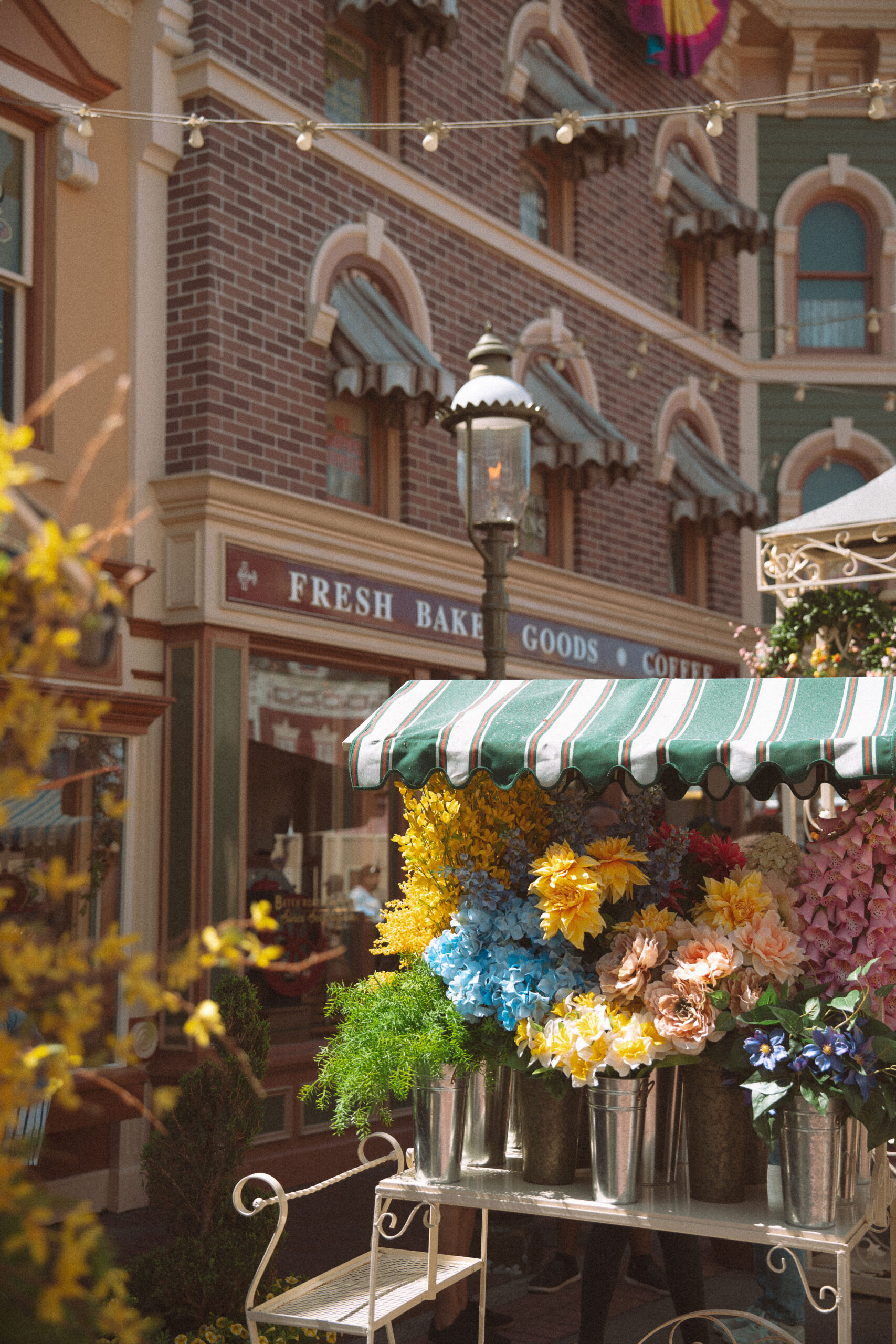 Colorful flower market stall with yellow, blue, and pink blooms on Main Street USA at Disneyland, with string lights and a green striped awning in warm morning light