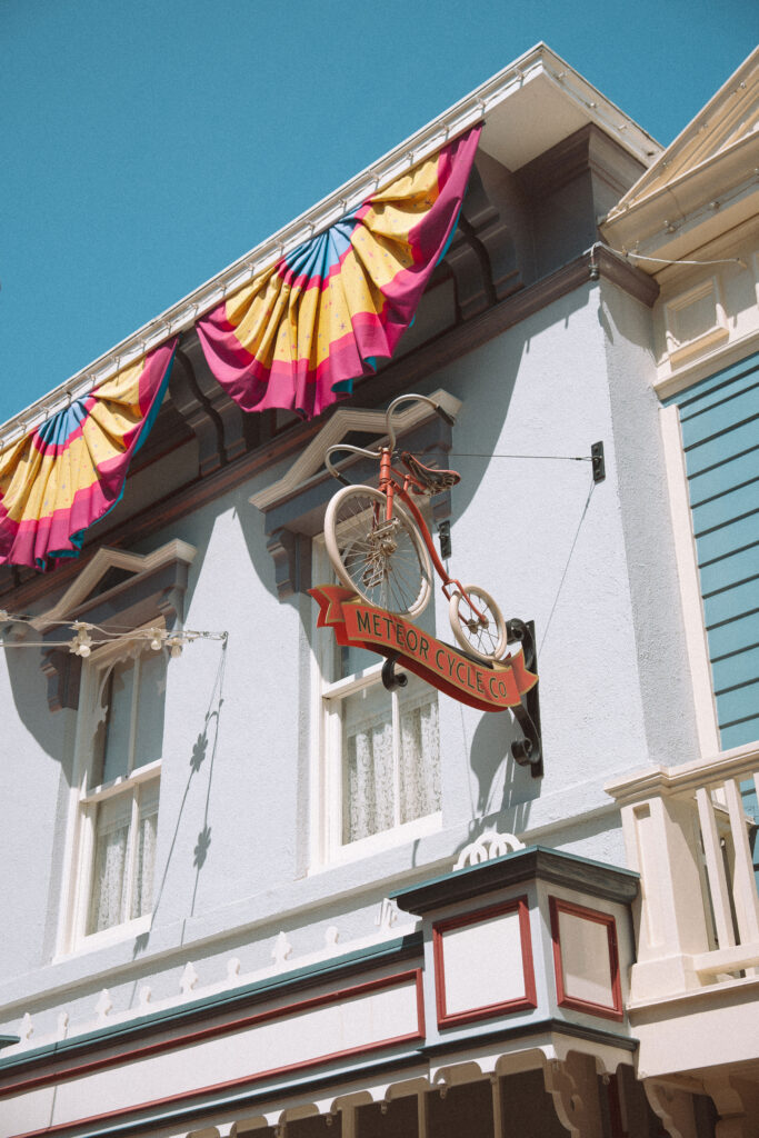 Vintage penny-farthing bicycle sign for Meteor Cycle Co. on Main Street USA at Disneyland, with colorful pink and yellow bunting against a bright blue sky