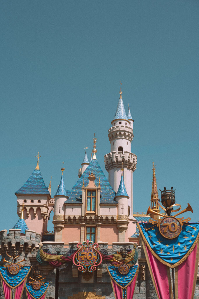 Sleeping Beauty Castle at Disneyland decorated with 70th anniversary blue and pink banners and gold fanfare ornaments against a clear blue sky