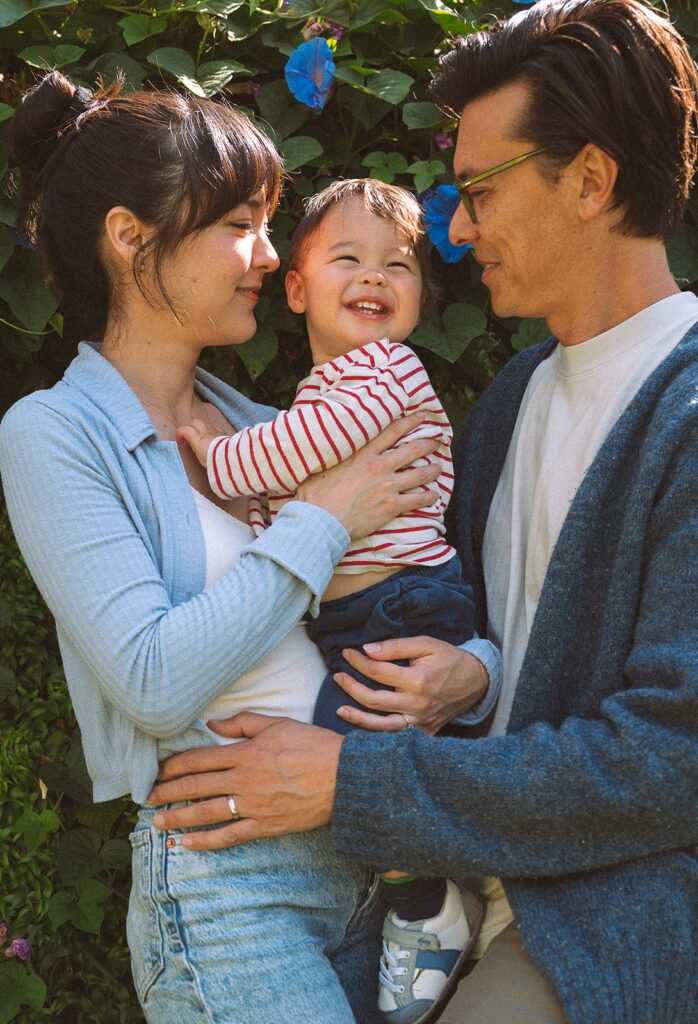 Parents holding their laughing toddler between them outdoors during a lifestyle family session in Long Beach, morning glory vines in the background