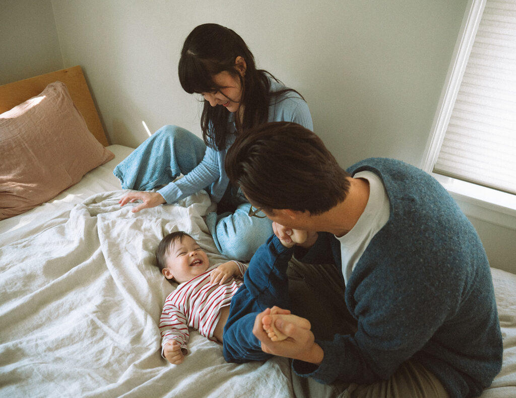 Candid at-home lifestyle family session with parents playing with their giggling toddler on the bed in soft morning window light