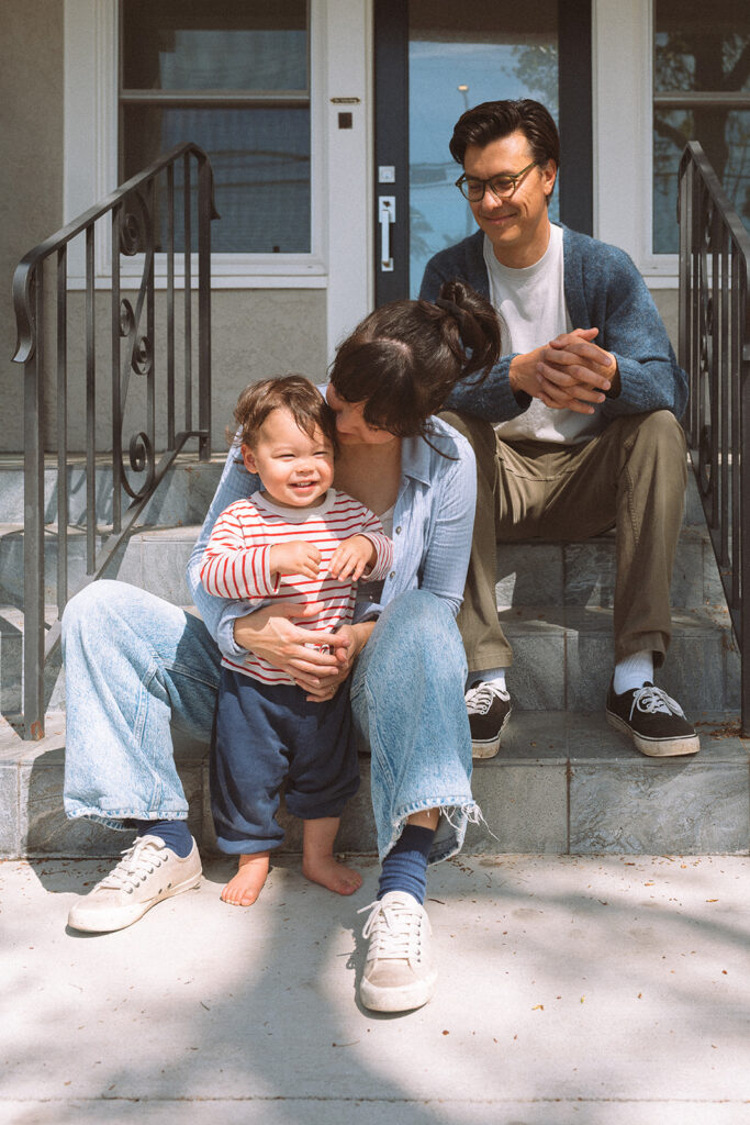Family of three sitting on front porch steps during a lifestyle family session in Long Beach, mother kissing toddler on the cheek while father smiles behind them
