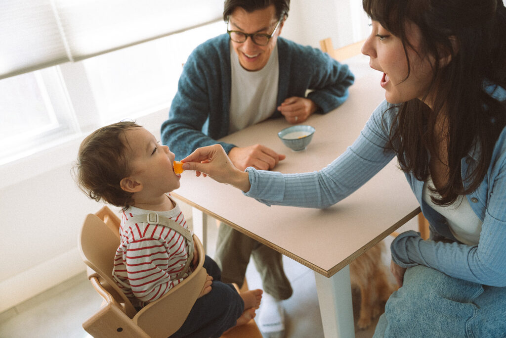 Father laughing while mother feeds toddler an orange slice in a high chair during a candid family mealtime photo