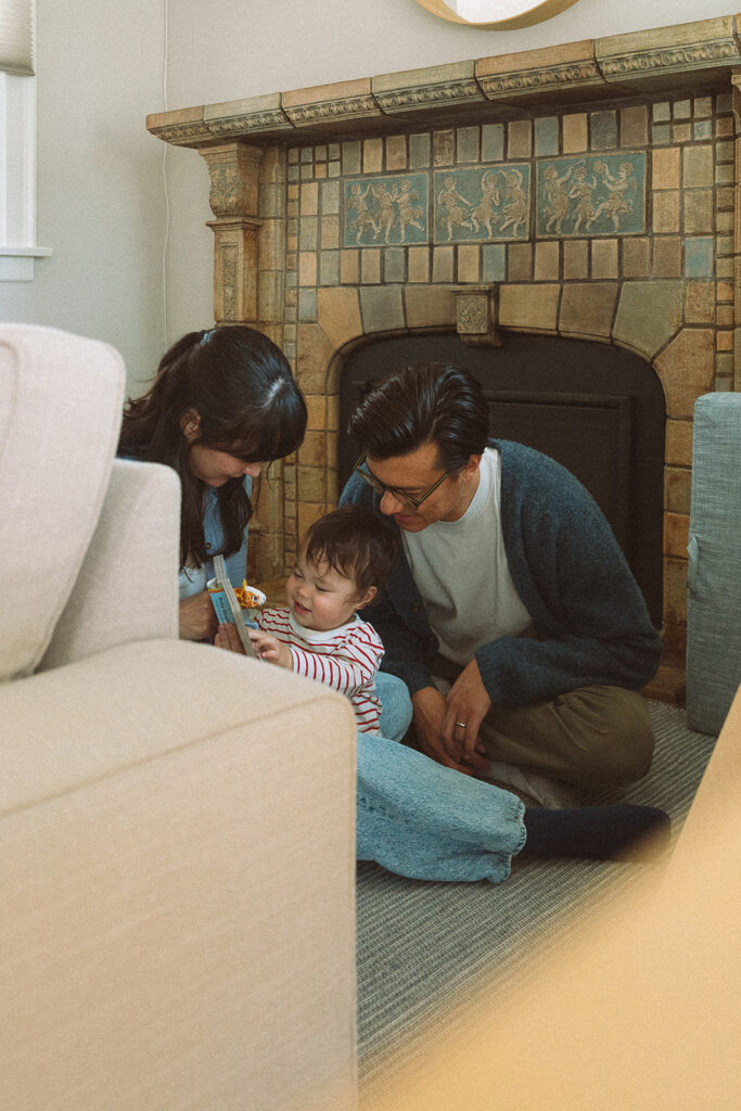 At-home lifestyle family session in Long Beach, parents and toddler reading together on the floor in front of an ornate fireplace
