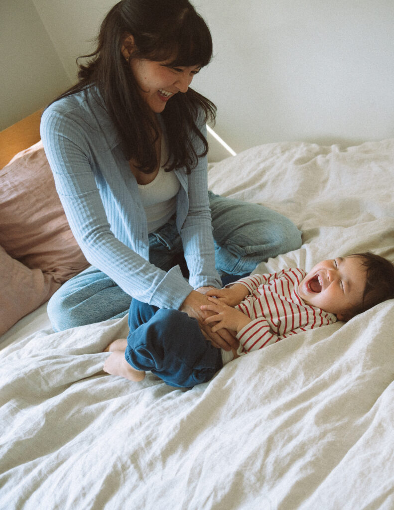 Mother laughing and tickling her toddler on the bed, candid moment captured during an at-home documentary family session