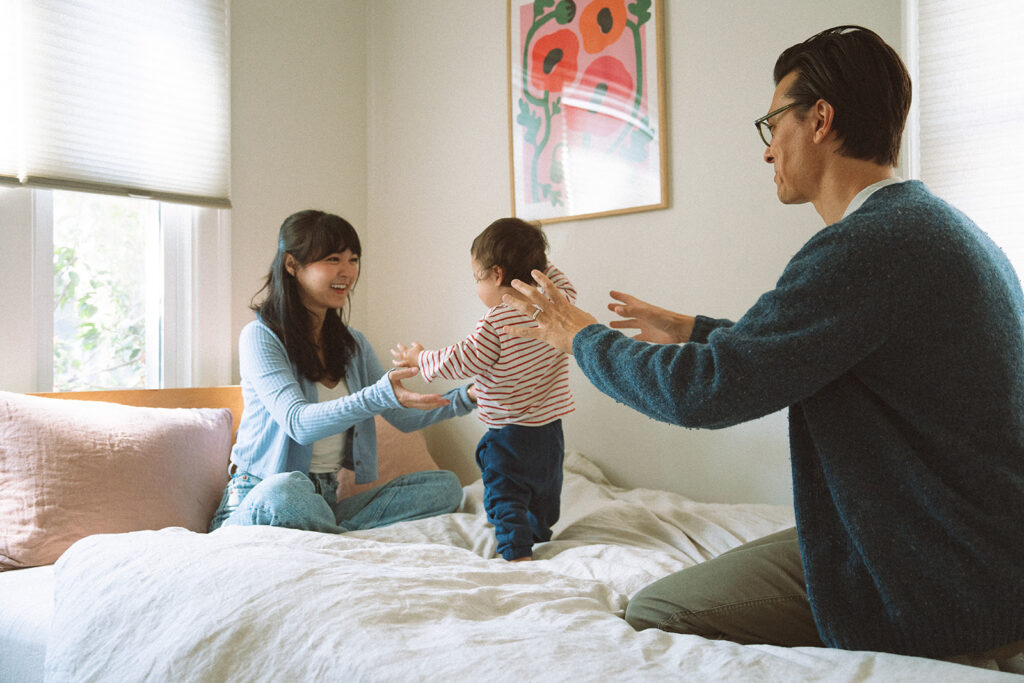 Toddler taking first steps on the bed between both parents during an in-home family session, colorful art print on the wall behind them