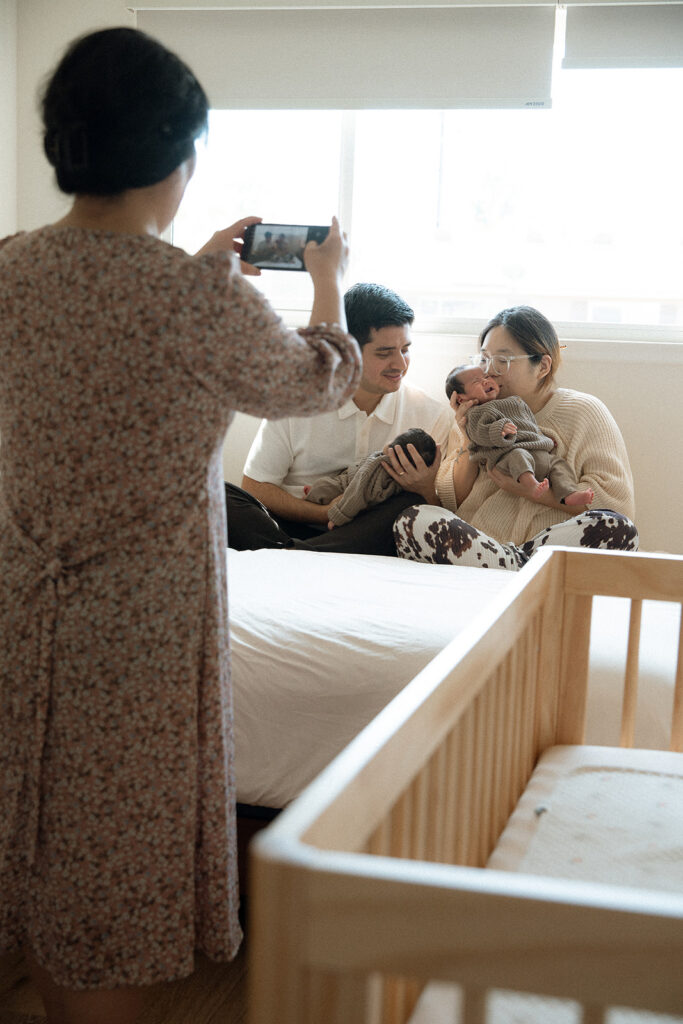 Grandmother photographing new parents holding newborn twins on a bed in their Irvine home — Orange County in-home newborn photographer