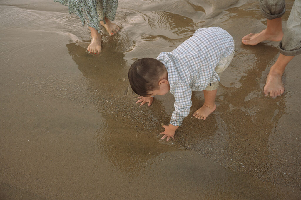Toddler exploring wet sand between his parents' bare feet at Crescent Bay Beach during a family photography session