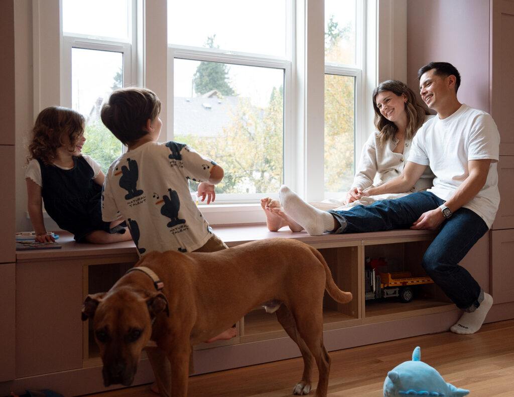 Parents sit on a pink window bench laughing with their two kids and family dog during a natural-light at-home family session in Orange County.