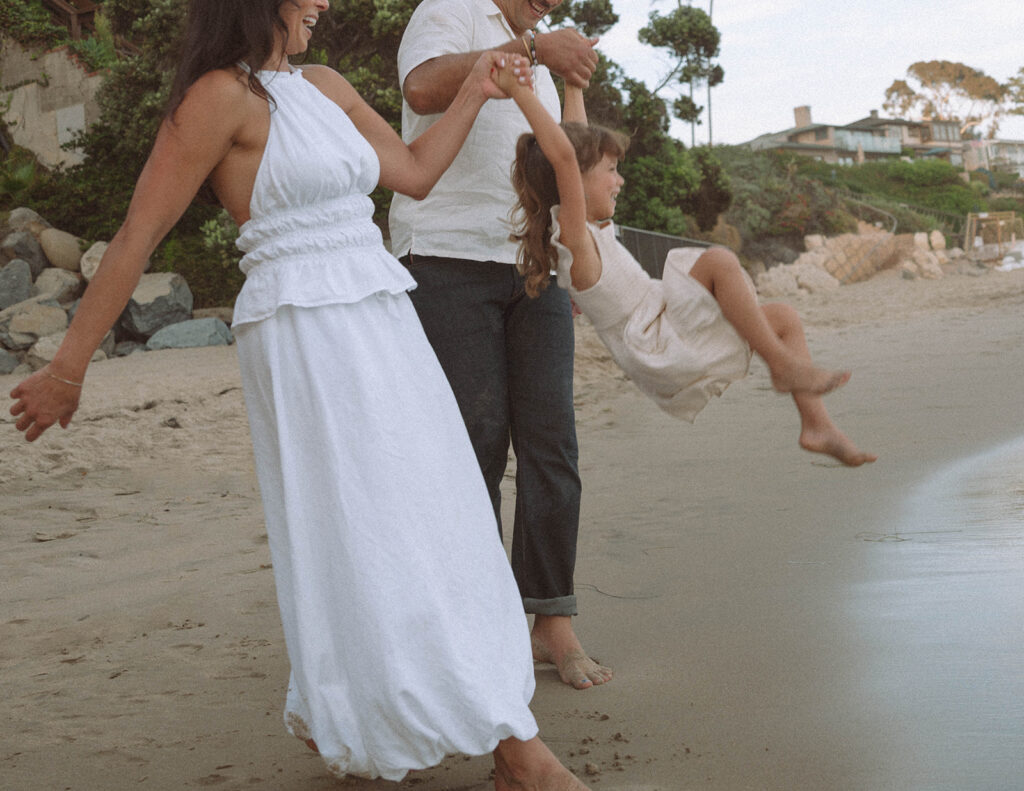 Parents swing their daughter between them along the waterline during a candid beach family session in Laguna Beach, Orange County.