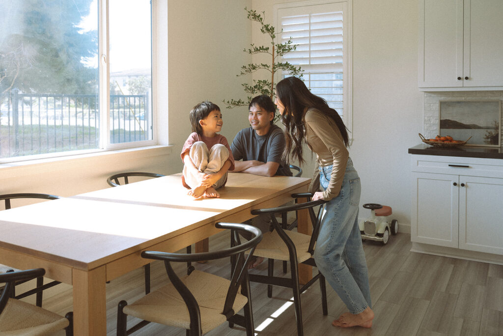A young boy sits cross-legged on the kitchen table chatting with his parents in soft window light during a documentary-style family session.