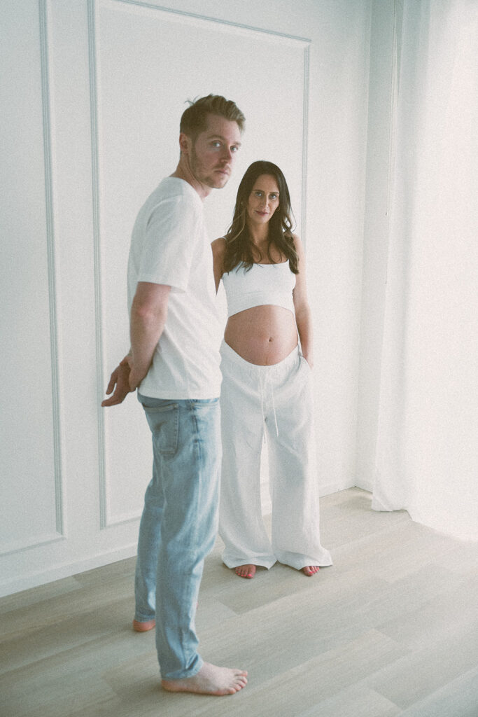 A couple stands close by a soft white wall, her baby bump cradled between them. Studio family sessions in Orange County.