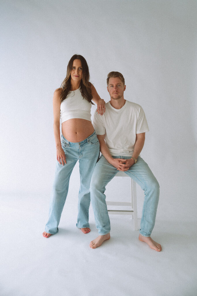 Parents-to-be in white and denim, her bump showing, him on a stool in the studio.