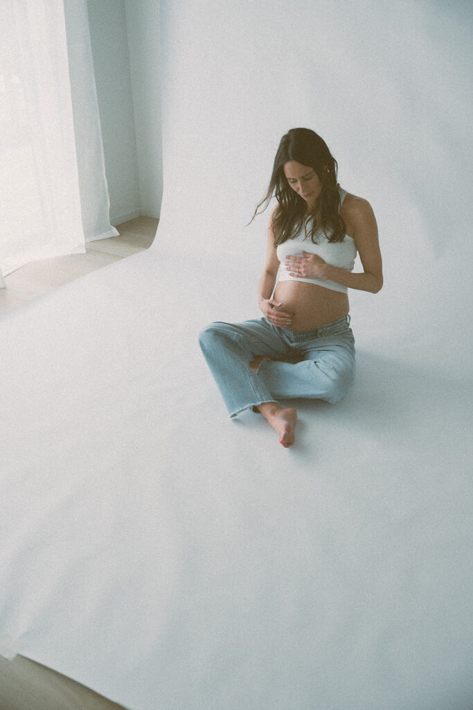 A pregnant woman sits cross-legged on the studio floor, both hands resting on her belly.