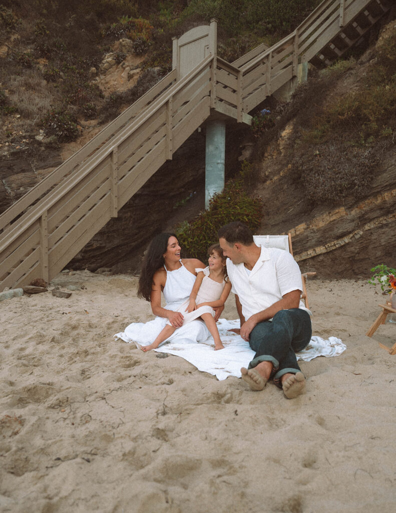 Parents sit on a blanket in the sand with their young daughter during a relaxed beach family session at Crescent Bay, Laguna Beach.