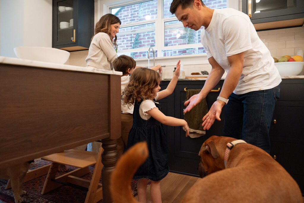 Two young siblings wash their hands at the kitchen sink while dad hands them a towel and the family dog watches from the foreground.
