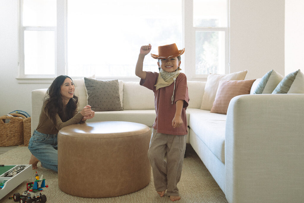 Young boy in a cowboy hat and bandana strikes a pose while his mom laughs behind an ottoman during an at-home family session in Orange County.