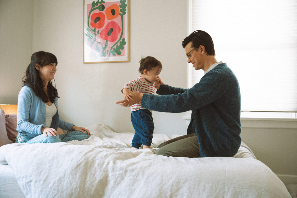 Dad helps a toddler in a striped shirt stand on the bed while mom watches and laughs, captured during an Orange County in-home family photoshoot.
