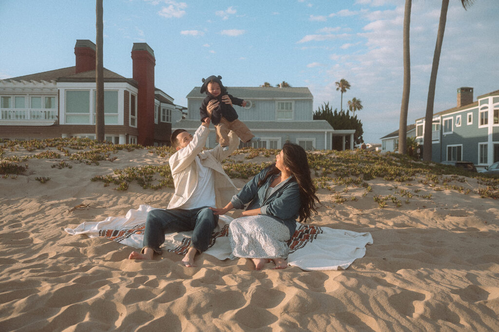 Dad lifting baby into the air while mom watches from a blanket on the sand, Newport Beach beach houses and palm trees in background.