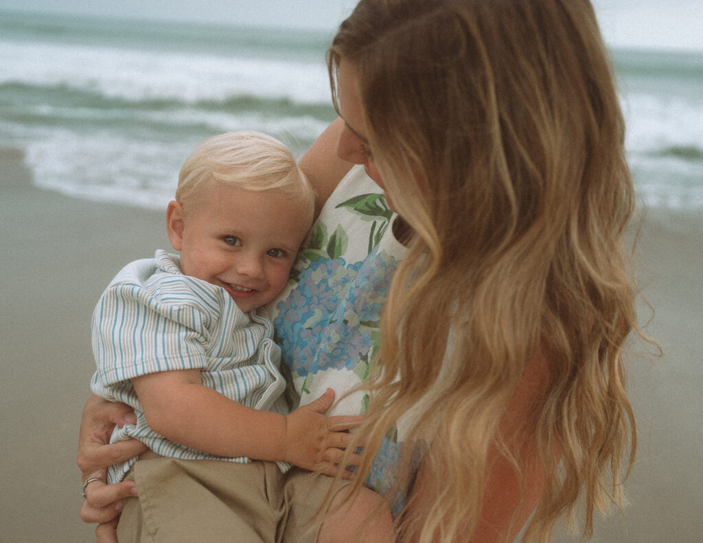 Toddler boy smiling over his shoulder while held close by his mom on the sand in California.