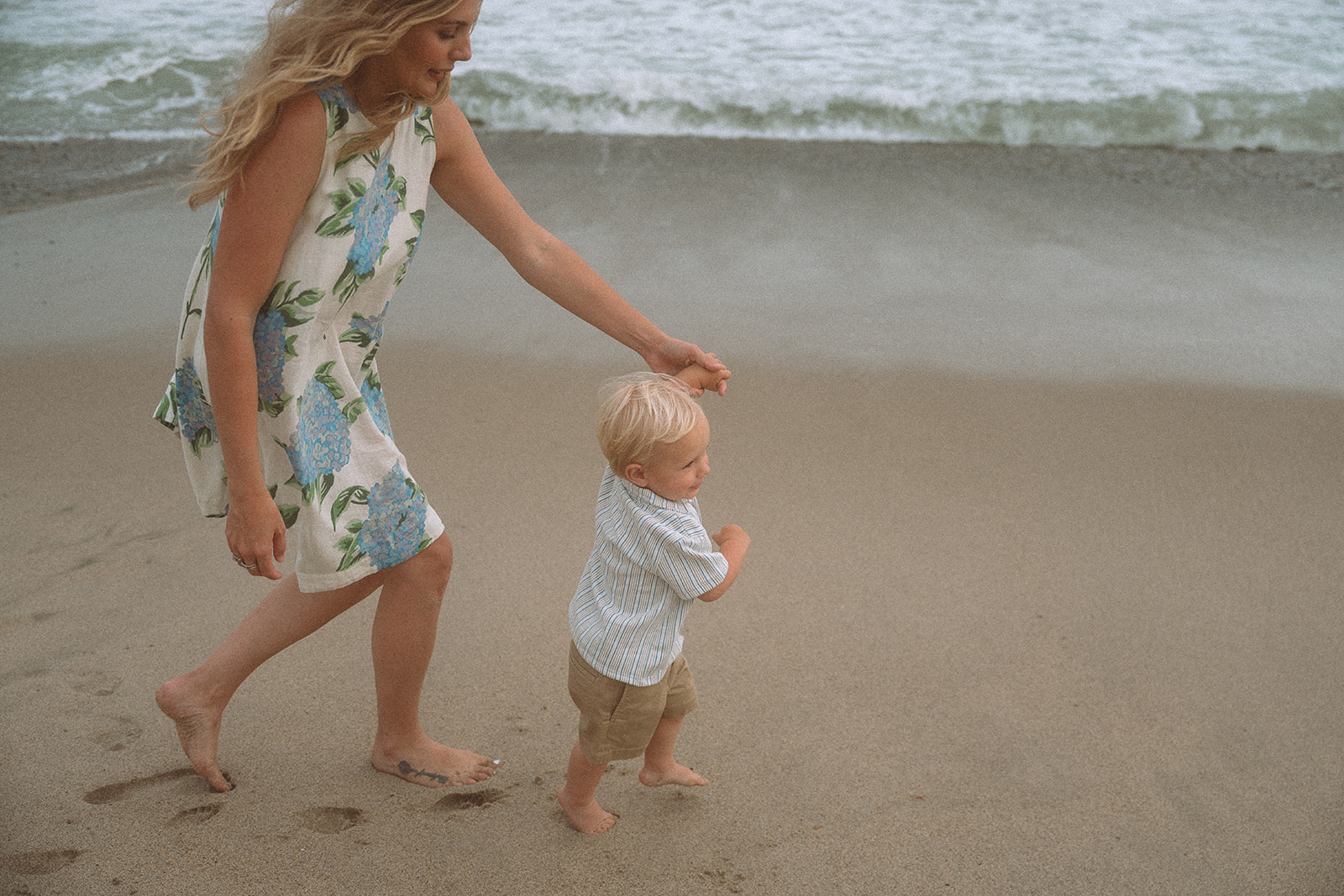 Mother and toddler son walking hand in hand along the sand, photographed by a Dana Point family photographer at Capistrano Beach