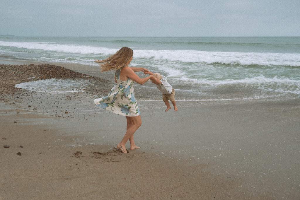 Mother swinging her toddler son by the arms on the shoreline, captured by an Orange County family photographer.