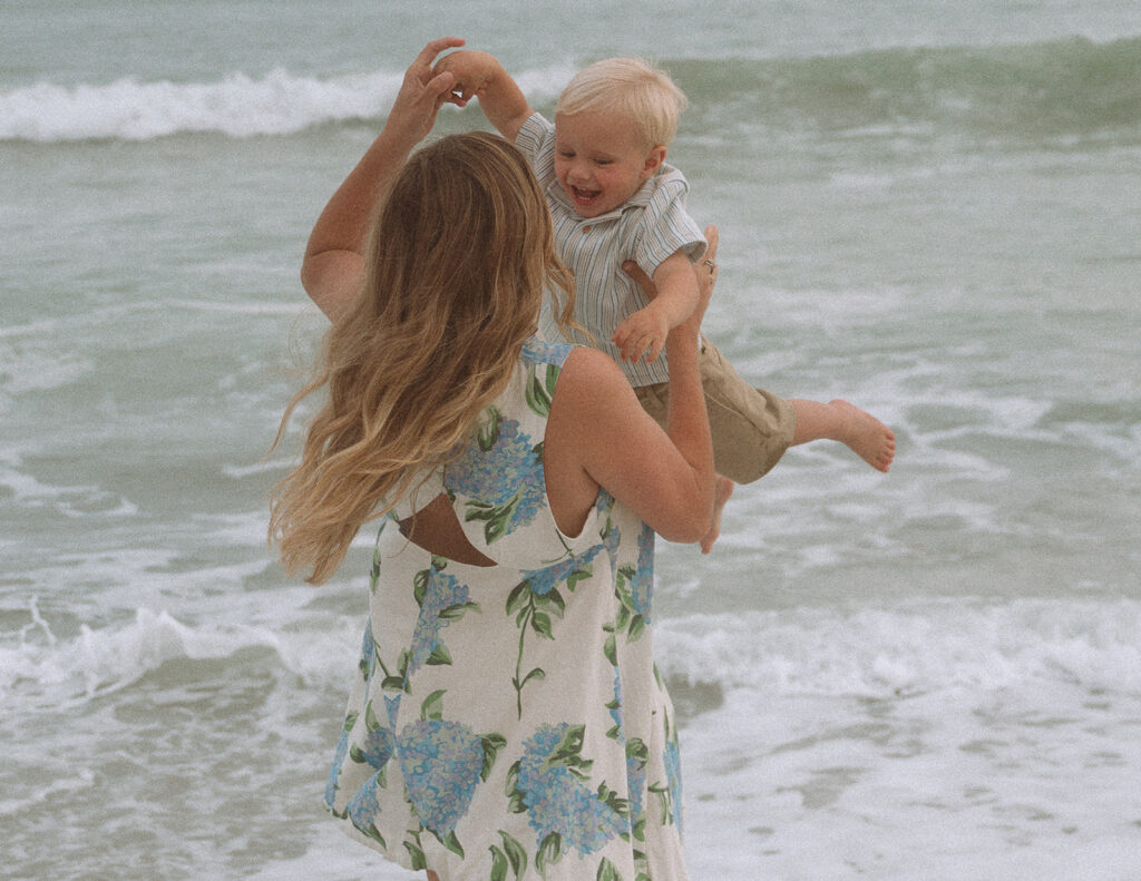 Mother lifting her laughing toddler son into the air at the water's edge, photographed by Maria Alcantara photography.