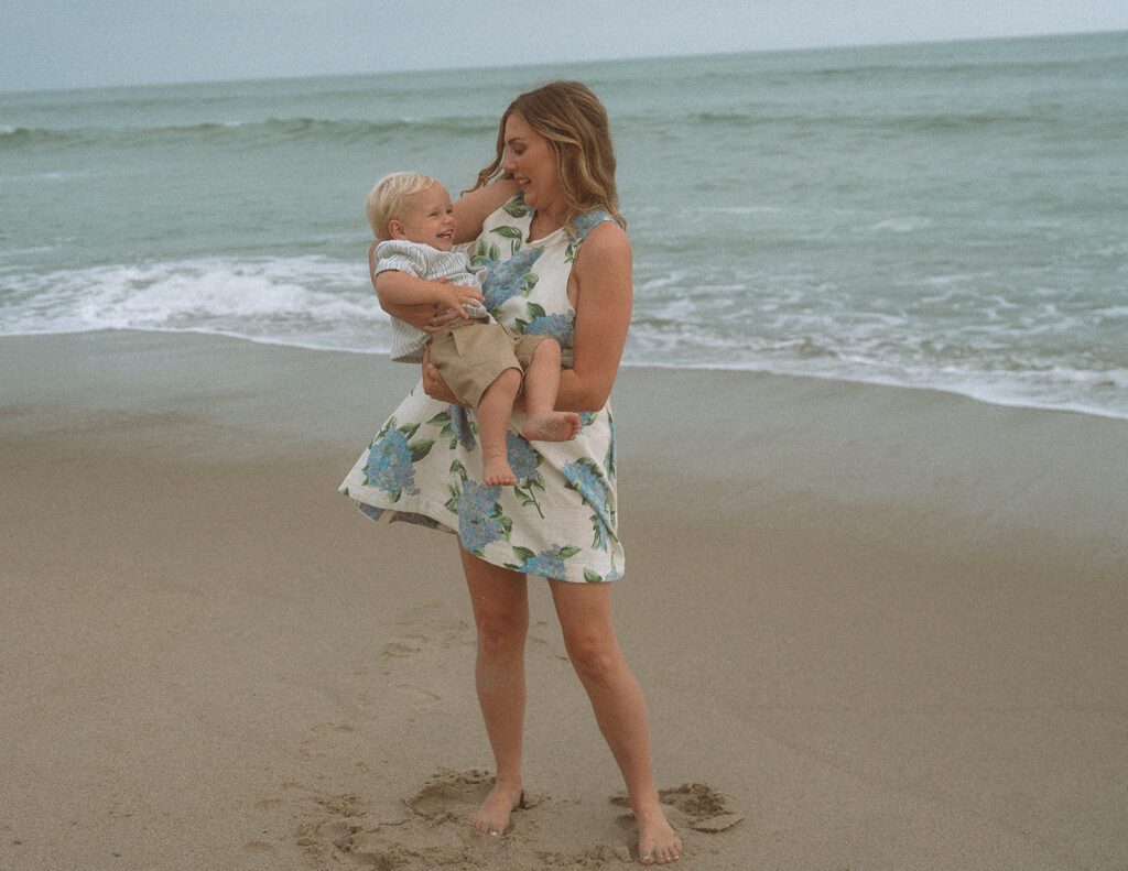 Mom in a blue floral dress holding her giggling toddler son, both barefoot on the beach at Capistrano Beach.