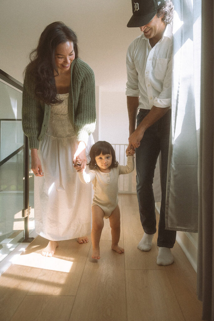 Toddler taking her first steps between mom and dad in a sunlit hallway.