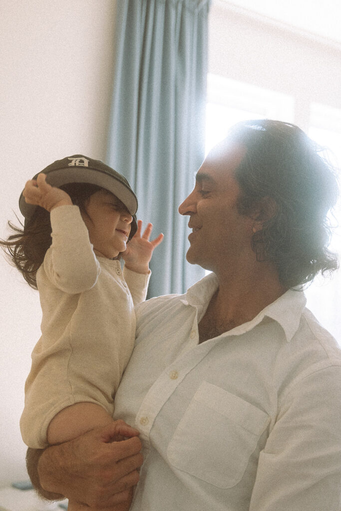Baby boy grabbing dad's baseball cap while he holds her up laughing.