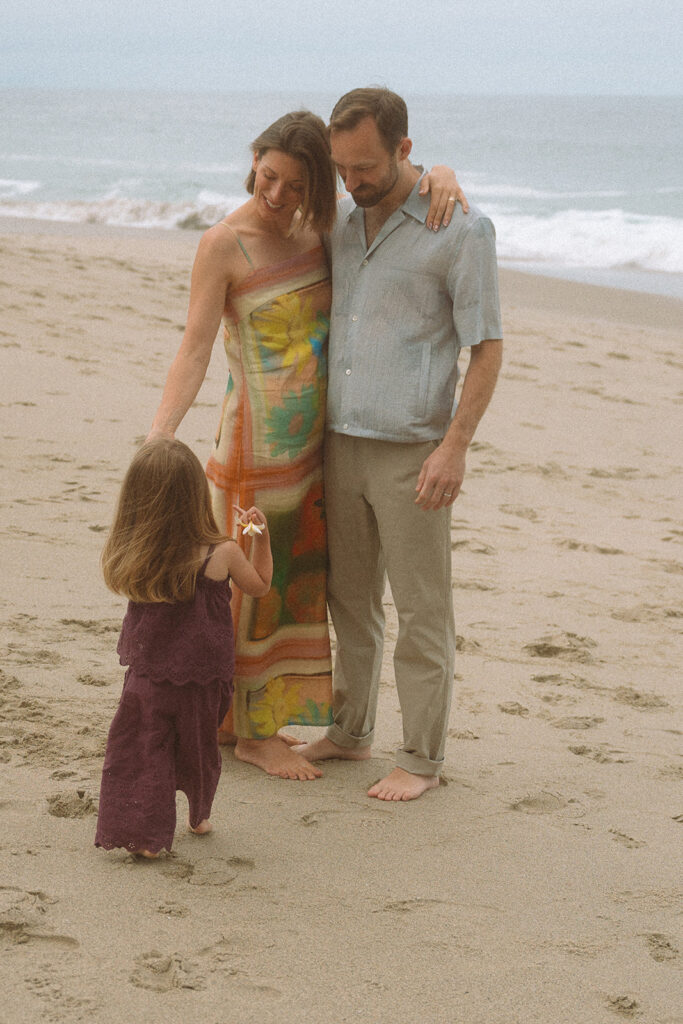 Mom in a colorful dress and dad walk the sand as their toddler reaches up during a Laguna Beach family session.