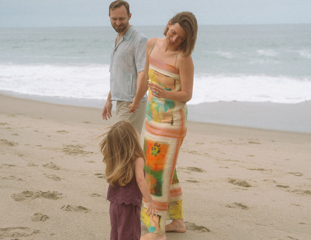 Parents embrace at the shoreline as their toddler holds up a flower during a Victoria Beach family session in Laguna.