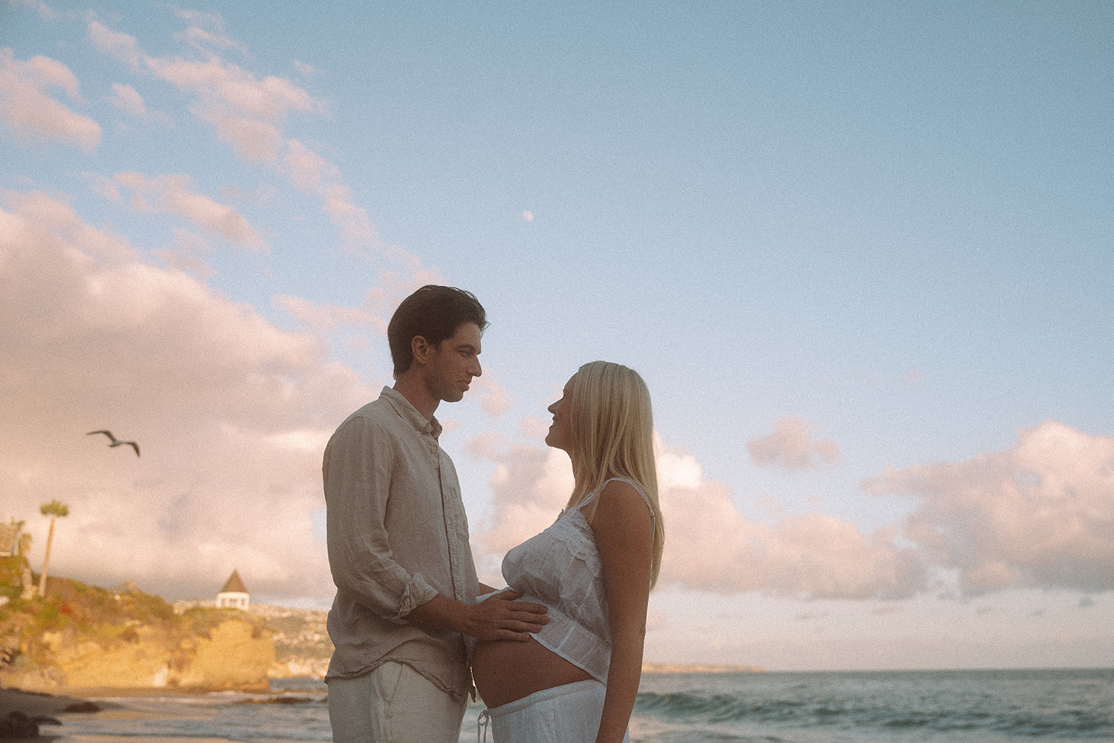 Expecting couple holding baby bump together on Shaw's Cove beach with dramatic pink clouds and ocean at golden hour, Laguna Beach maternity session.