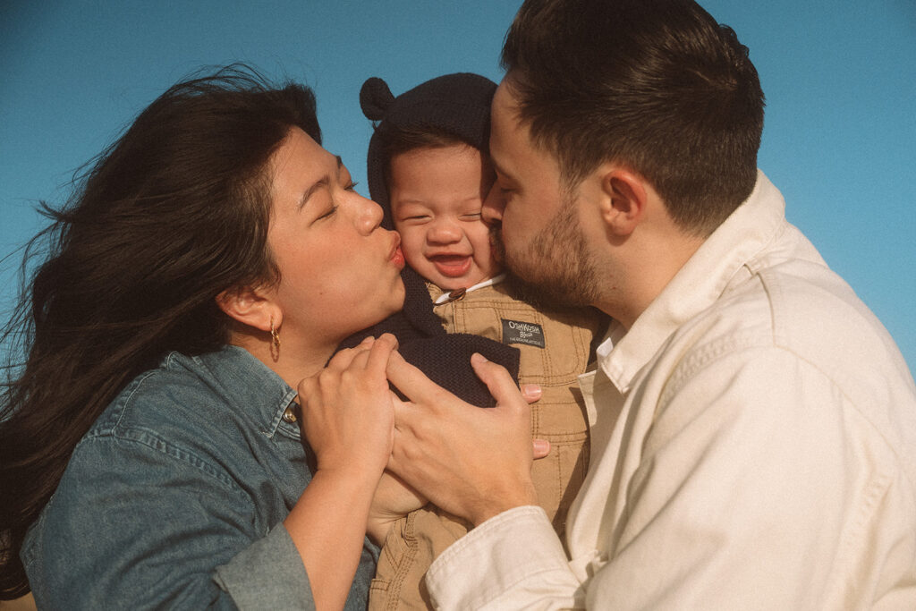 Newport Beach baby photographer captures mom and dad kissing their laughing baby boy on each cheek against a bright blue sky.