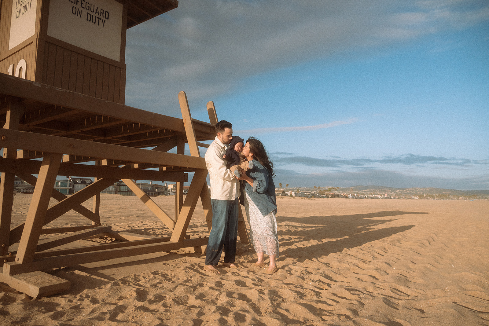 Family of three standing together by a lifeguard tower during a lifestyle beach session, golden afternoon light.
