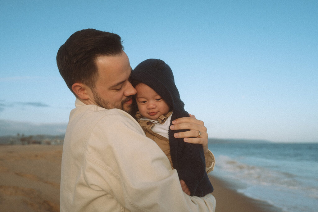 Dad holding baby close and nuzzling his head at dusk, ocean waves in the background, Orange County lifestyle photography.