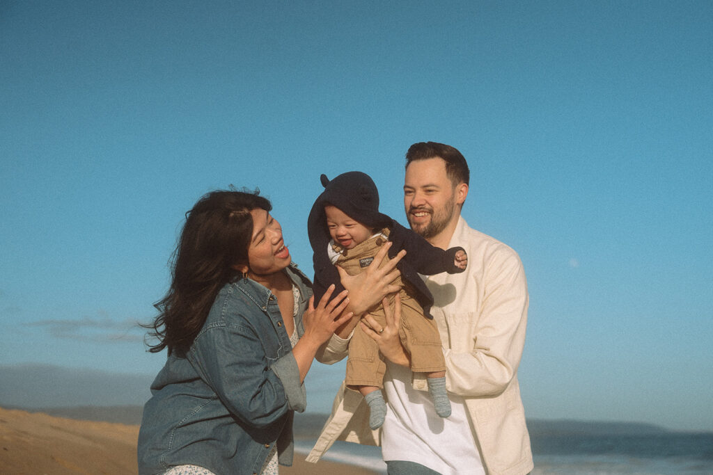 Dad holds baby boy while mom laughs beside them during a family photo session, clear blue sky background.