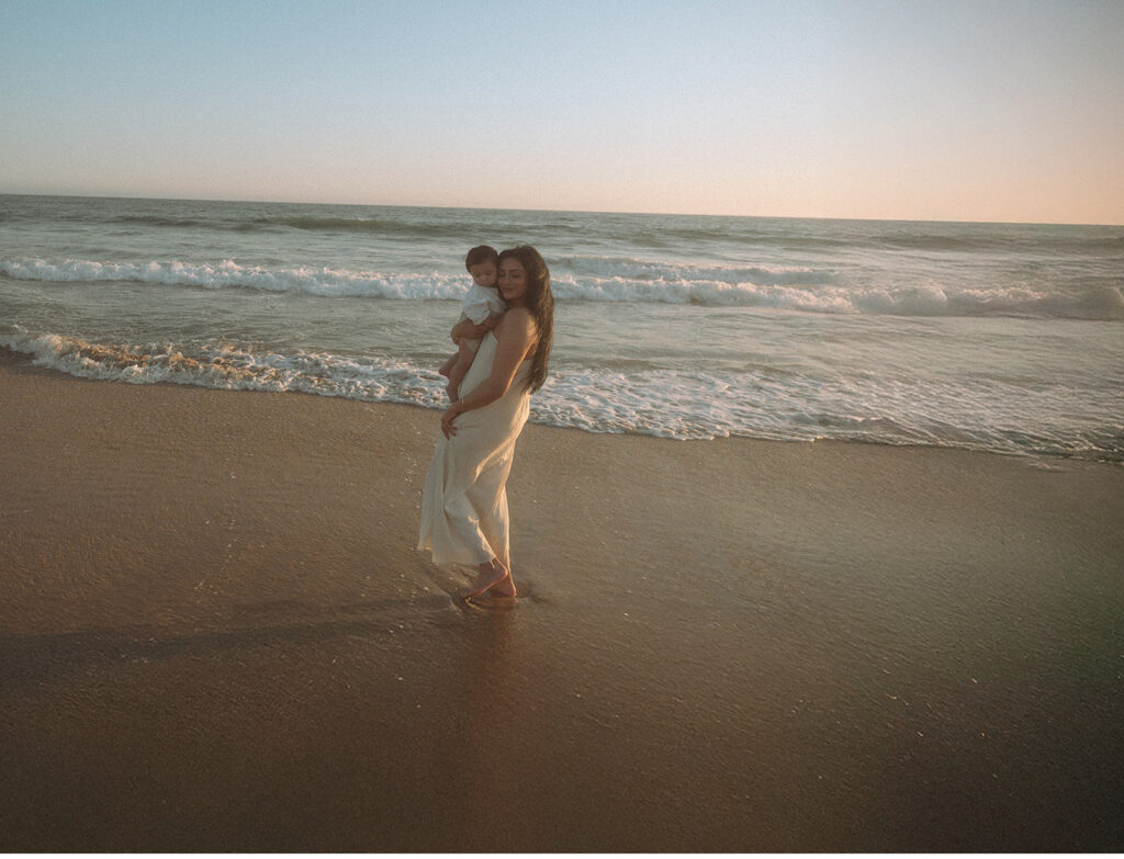 Mom in cream linen dress holding baby at the water's edge during Newport Beach sunset family session.