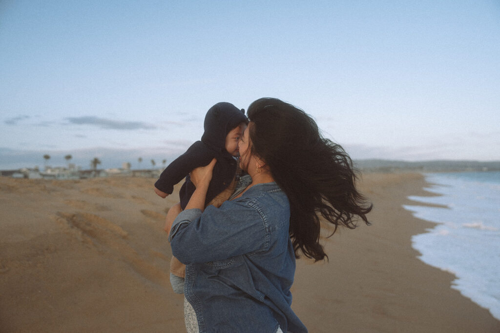 Mom lifting her baby boy toward her face at golden hour, hair blowing in the wind, Newport Beach shoreline at dusk.