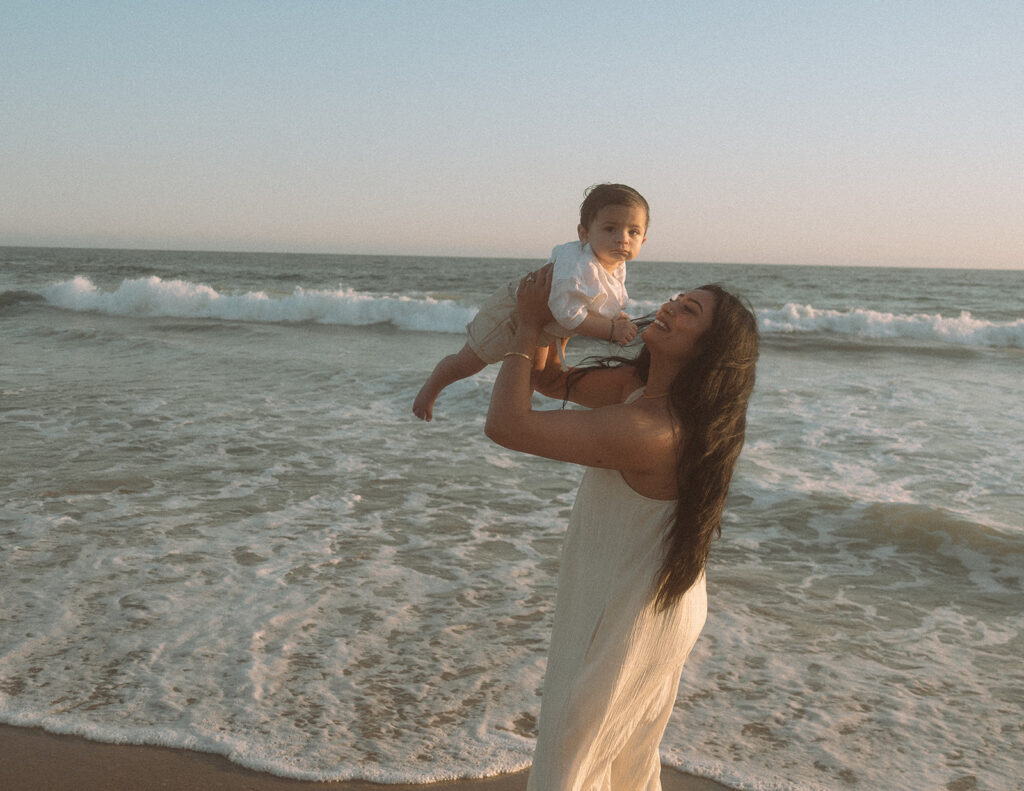 Mom laughing while lifting baby in the air with ocean waves behind them at Newport Beach.
