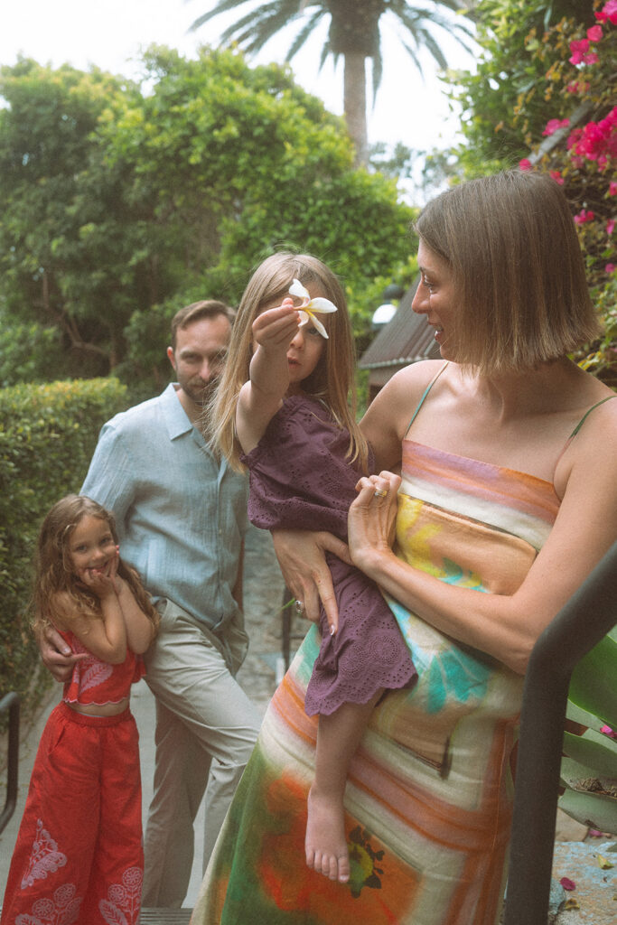 Mom holds her daughter, who lifts a plumeria flower, on a lush garden path during a Victoria Beach family session.