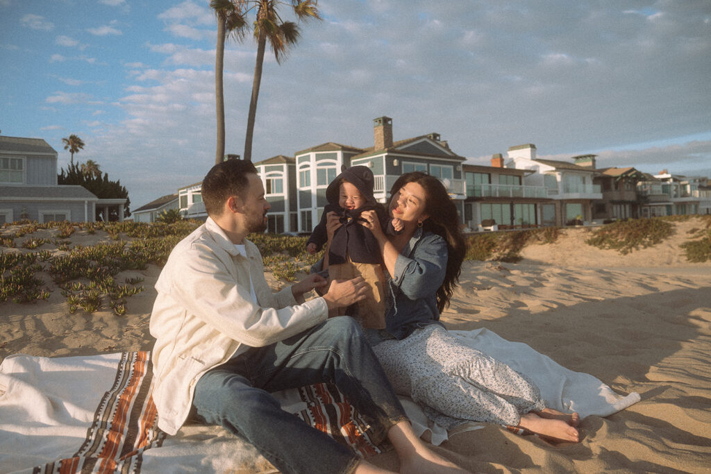 Newport Beach baby photographer — family sitting together on a blanket in the sand at golden hour, beach houses behind them.