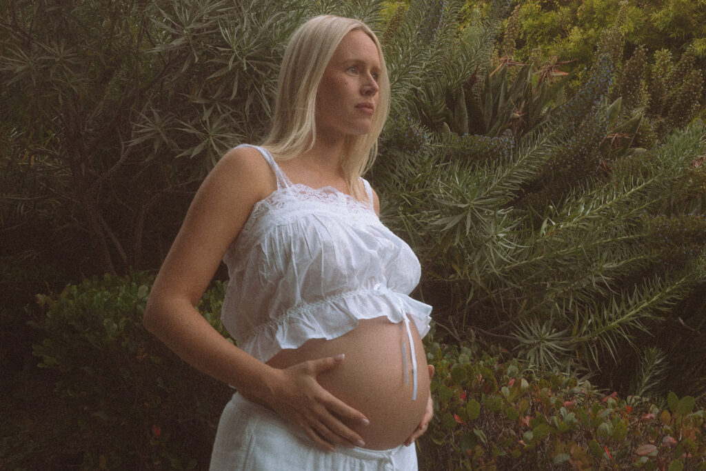 Pregnant woman in white lace crop top cradling her belly among lush coastal greenery in Laguna Beach, maternity portrait by Laguna Beach maternity photographer Maria Alcantara.
