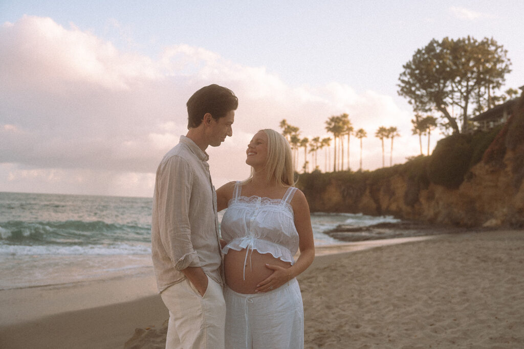 Maternity couple smiling at each other on Shaw's Cove beach at sunset with palm trees and cliffs in background, Laguna Beach.