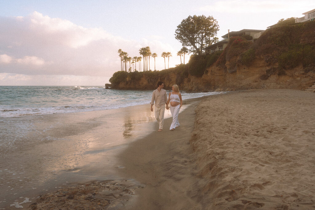 Expecting couple walking hand in hand along Shaw's Cove beach at golden hour, Laguna Beach maternity photographer.