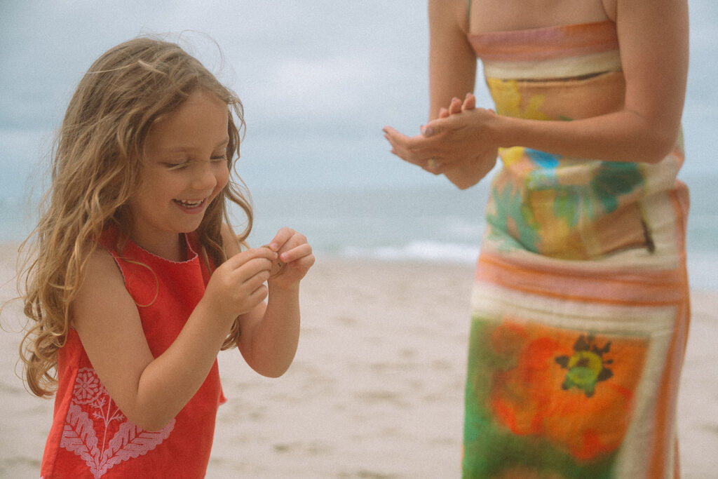 Young girl in red grins at a tiny shell in her hands while her mom stands nearby.