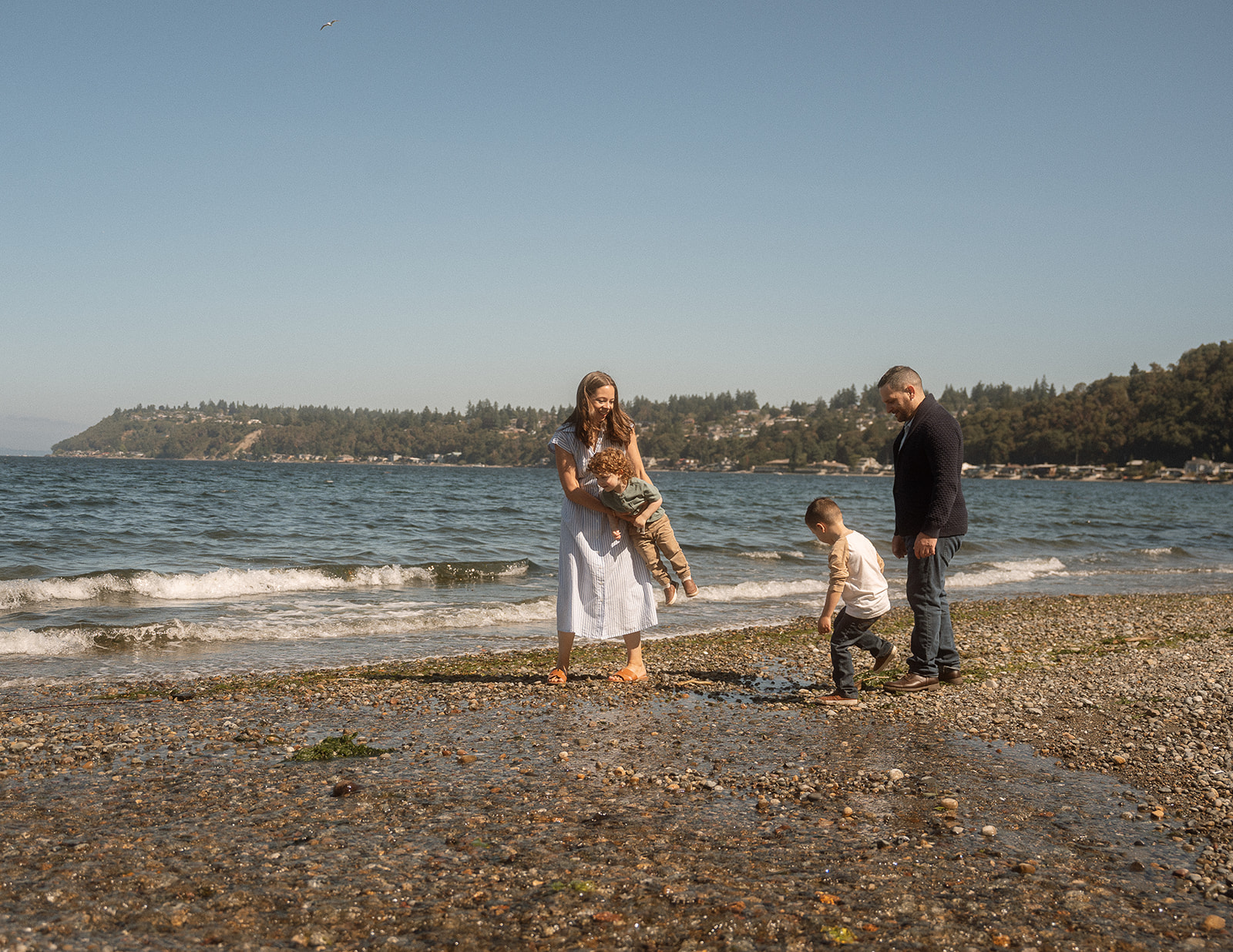 Family with two children exploring beach together with forested shoreline backdrop - natural playful family photos moment