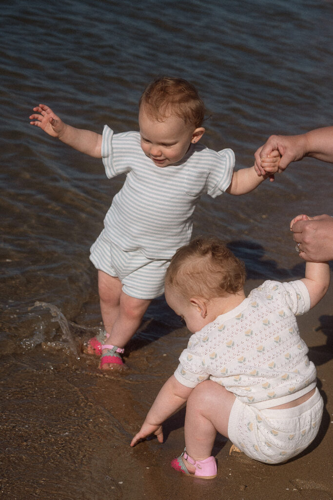 Toddler walking into a shallow beach holding a parent's hand as her sibling plays in the sand nearby.